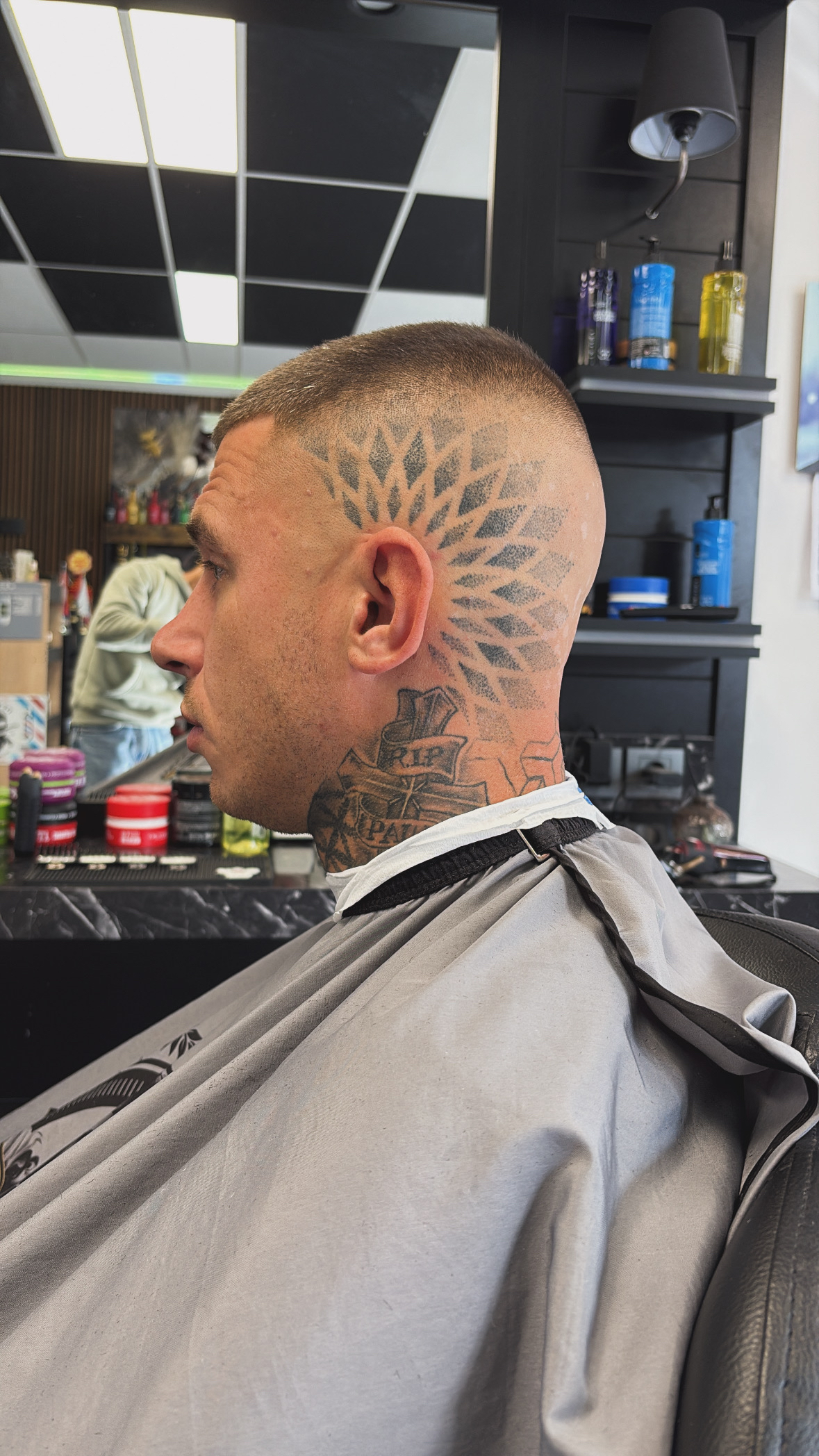 Klant met een kaalgeschoren fade rond een hoofdtattoo, met de schappen en het zwarte plafond van de zaak op de achtergrond